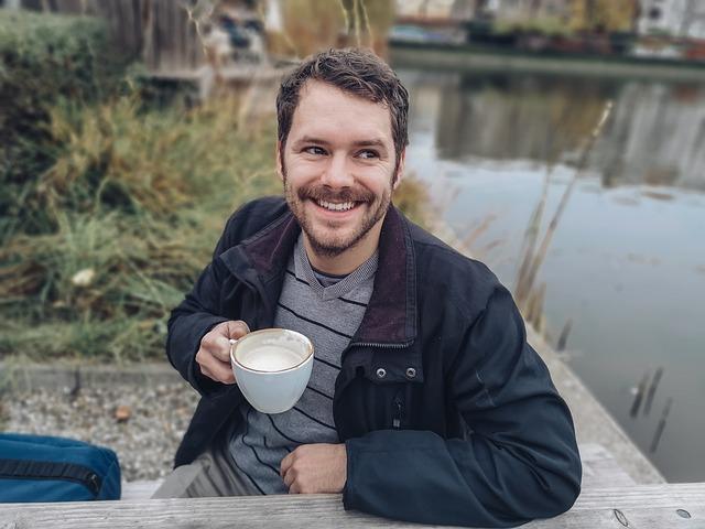 A happy man outdoors, sitting at a picnic table with a hot beverage. His coat is on and he is smiling.