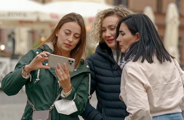 Three young women standing outside looking at something on a phone.