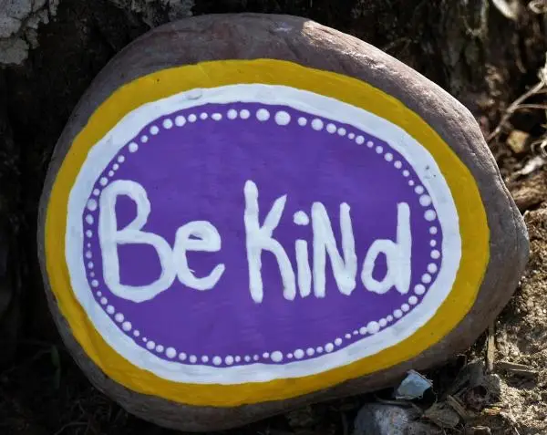 The words Be Kind on a round rock painted in purple and white with a yellow frame.
