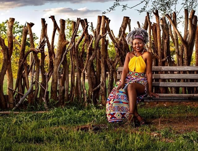 A young African woman wearing bright yellow sitting on a bench in a grassy space.