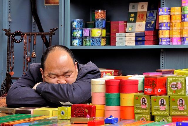 A man sitting at a table covered in items to sell, asleep with his head on his arms.