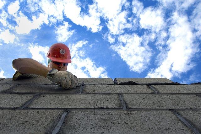 Looking up a cinder block wall toward the man with trowel and hard hat applying the mortar to build the wall.