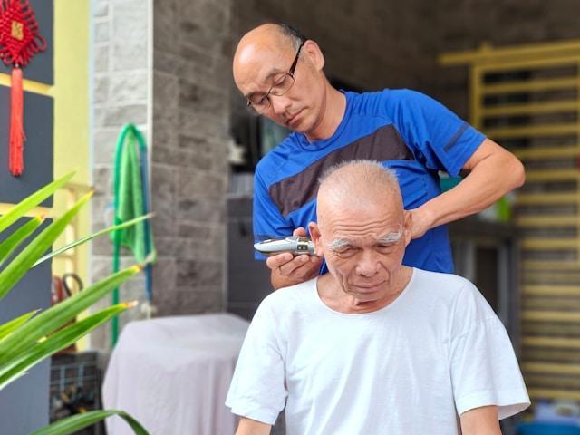 A bald man with glasses running a razor over the head of another elderly bald man.