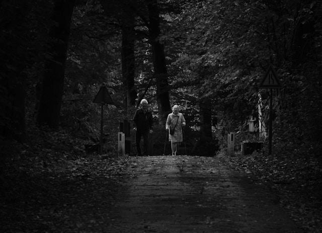 A black and white photo of an elderly couple walking along a dirt road through a forest.