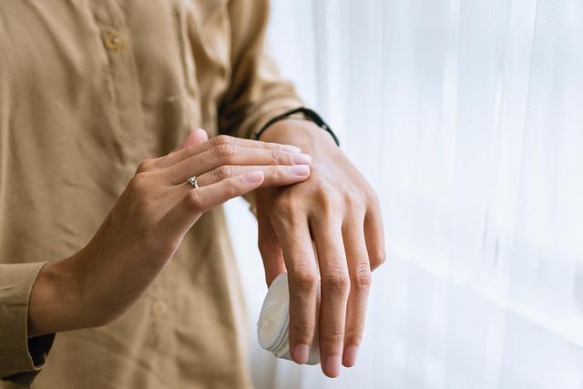 A close up of a woman's hands, one holding a small jar of cream, the other rubbing the back of her hand.
