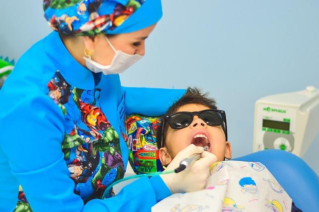 Female dentist dressed in bright blue scrubs, looking in the mouth of a teenaged boy.