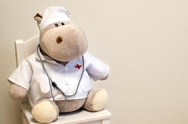 A beige stuffy hippopotamus dressed as a doctor with a stethoscope sitting on a child-sized chair.
