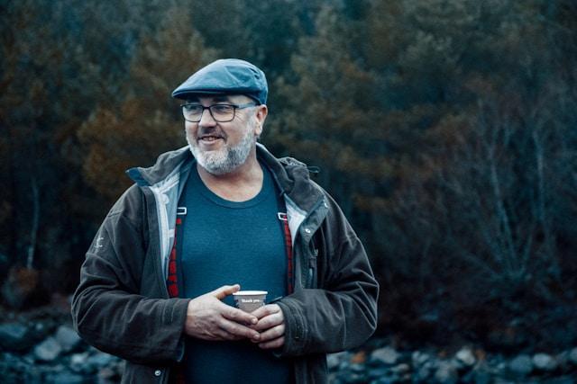An older man standing outside in the wilderness holding a cup of coffee.