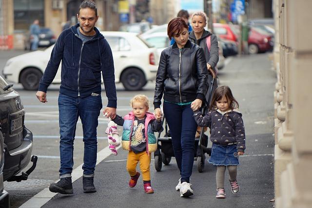 A family of 4 with 2 young children walking along a city sidewalk holding hands.