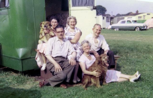 A photo from the 1950s showing an extended family of 5 with their 2 dogs outside their camper trailer.