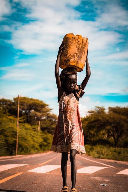 An African girl carrying a large container on her head walking down a paved road.