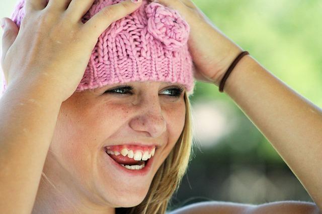 A girl with a pink knitted hat, hands on head, looking off to the side and laughing.