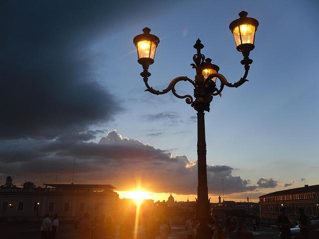 A lit three fixture lamppost in the city or Rome as the sun sets.
