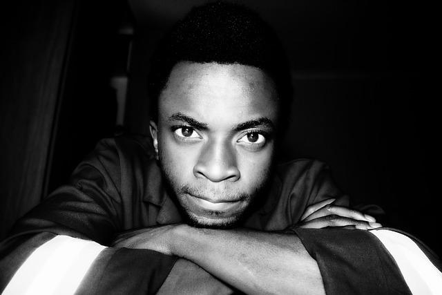 A close-up black and white image of a black man, arms folded staring directly at us.