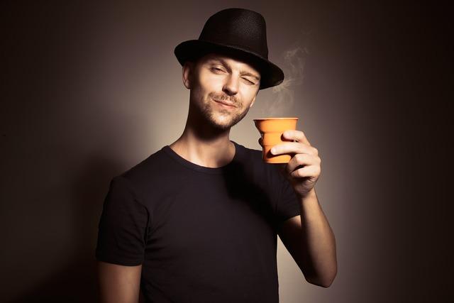 A man with a bowler hat, scruffy beard and wink, holding up a small cup of coffee.
