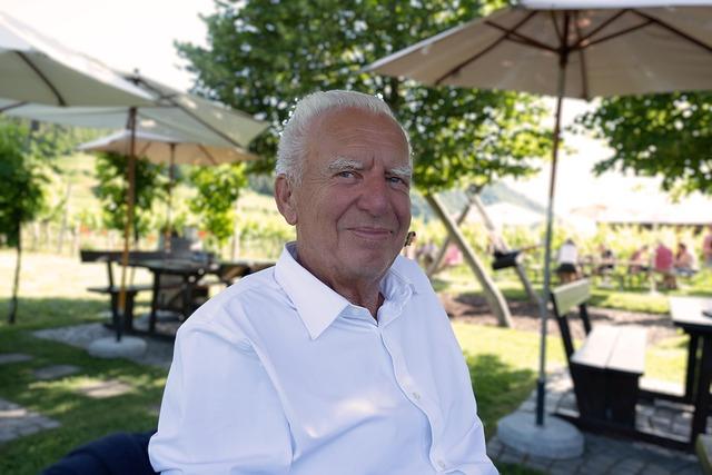 An old man with a white shirt, looking directly at us with a small smile, sitting in an outdoor picnic area in the shade.