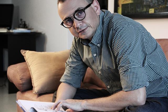 Man with glasses sitting forward on a couch with his elbows on his knees holding a book and looking at the camera.