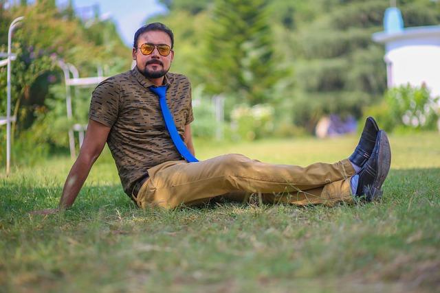 A young Asian man with glasses, nice clothes and a blue tie sitting with legs out straight on the grass.