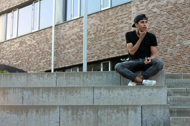 A young man wearing a baseball cap backwards, sitting on a concrete wall looking out toward his left.
