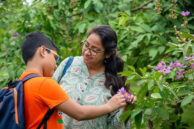 A brown-skinned mom and pre-teen son, both wearing glasses standing by a flowering shrub. The son is looking at a flower and the mom is looking at the son.