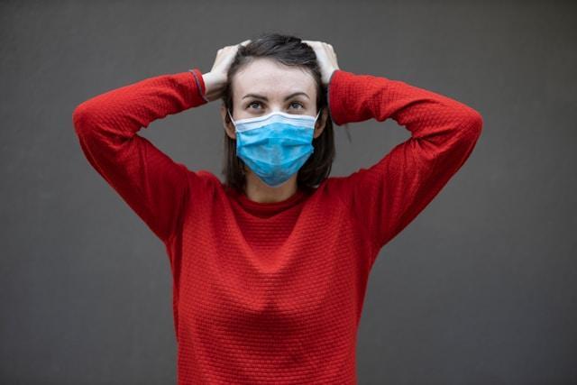 Woman in a red long-sleeved top wearing a blue mask. She has her hands on her head and is looking upward.