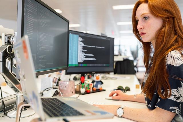A side view of a woman in an office at a desk looking at one of her 3 computer screens.