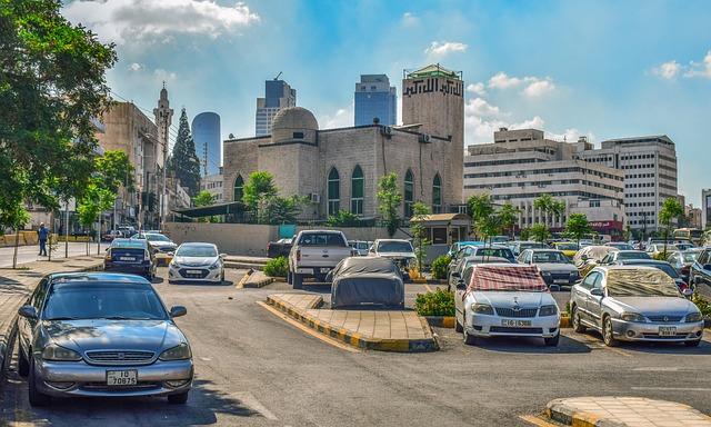 An urban parking lot with several cars in the lot and a cityscape behind.