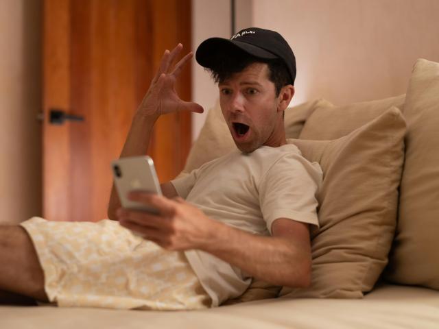 A young man, baseball cap & t-shirt, lounging on a bed, phone in hand, other hand raised and an expression of disbelief.