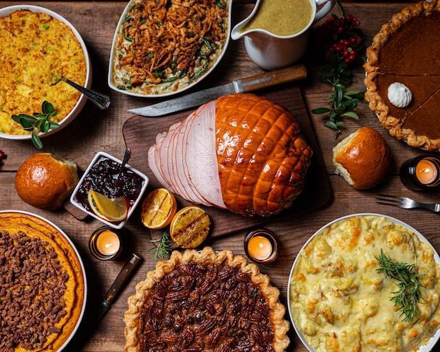 a close-up image of roast ham, pecan pie, pumpkin pie, mashed potatoes and other food from a Thanksgiving meal