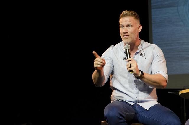 A middle-aged man with mic in hand sitting on a stool presumably speaking to an audience.
