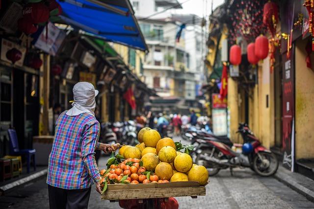 A man with a cart full of melons and oranges walking down a narrow street.
