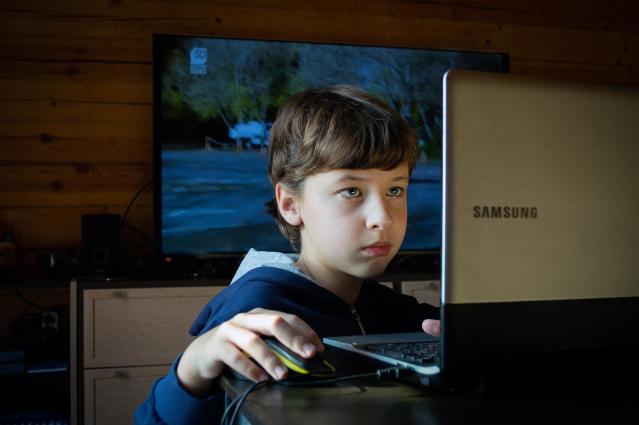A young teen holding the mouse and looking at his computer screen.