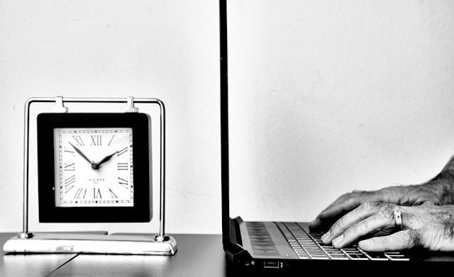 A black and white image of a laptop and an analogue clock showing 8 minutes to 2.