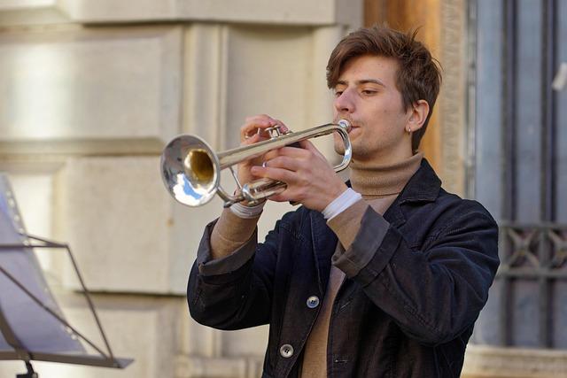 A young man in jacket and turtleneck playing a silver trumpet outdoors.