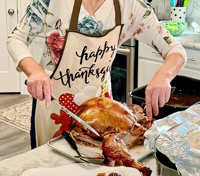 A woman carving a turkey wearing an apron that reads 'happy thanksgiving' in cursive.