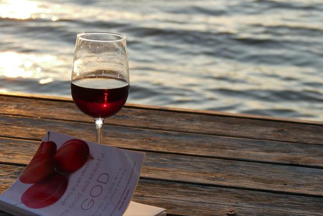 A glass of red wine and a book on a dock by a lake.