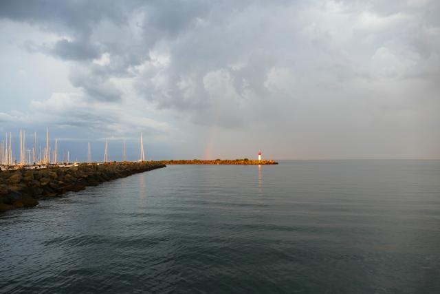 A photo from shore looking out along a long breakwater to a navigational marker on the end of another breakwater with a rainbow and dark clouds behind.