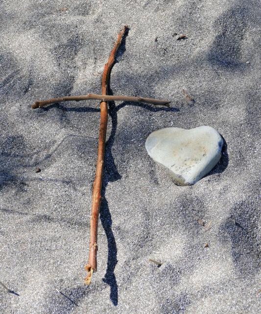 Two small twigs in the shape of a cross with a heart-shaped rock to the right on a sandy beach.