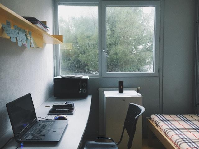 A wall-desk, single chair, open laptop and a small bed in a small room.