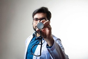 A young doctor with a white lab coat holding up a stethoscope and partially blocking his face.