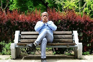 A man sitting on a park bench, legs crossed, covering his mouth and nose for a sneeze.