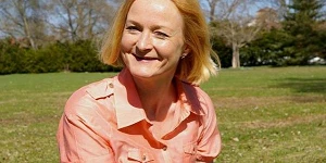 A woman with blond hair and an orange shirt sitting on grass on a sunny day, looking directly at us with a cheery smile.
