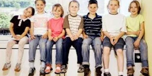 Seven children sitting on an indoor ledge, their feet dangling down.