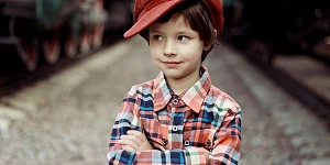 A young boy in a red cap askew on his head, a red & blue plaid shirt, arms crossed, looking to his right with an impish smile.