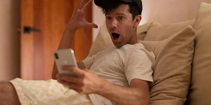 A young man, baseball cap & t-shirt, lounging on a bed, phone in hand, other hand raised and an expression of disbelief.