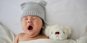 A baby yawning with a white teddy bear beside, covers pulled up to their chins.