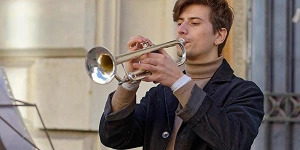 A young man in jacket and turtleneck playing a silver trumpet outdoors.