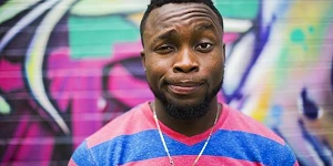A black man in a pink and blue striped t-shirt with a quizzical expression looking directly at us.