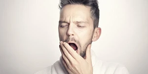 A young man, hair standing up, partially covering his mouth with his hand in the middle of a big yawn.