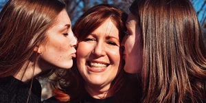 A mom with 2 grown daughters, one kissing each cheek while the mom smiles.